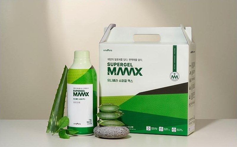 베이지 벽 앞 흰 바닥 위에 놓인 슈퍼겔 맥스 1 kg 병과 손잡이 달린 선물 상자, 알로에 잎, 쌓은 둥근 돌