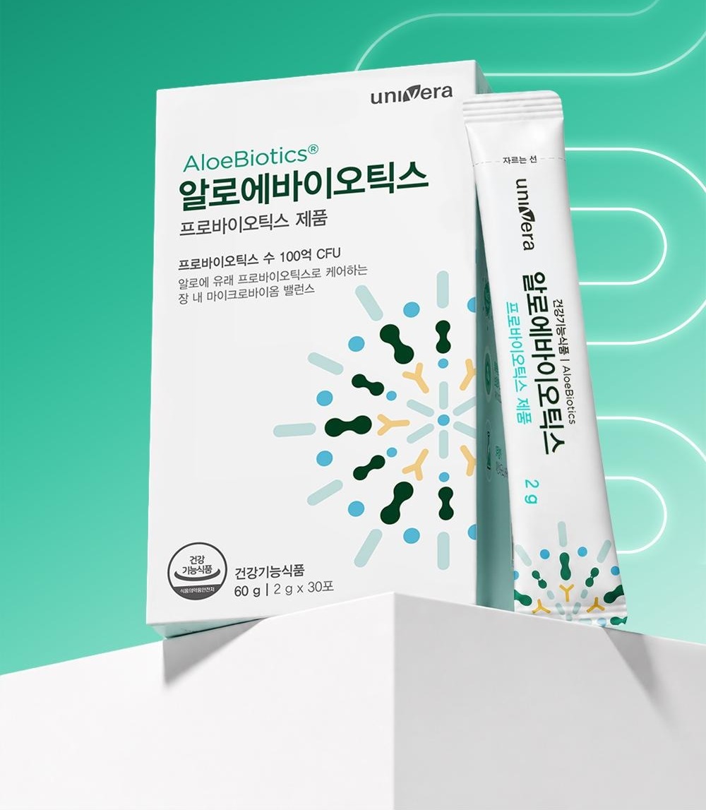 초록 그라데이션 지면 위 흰 단에 놓인 알로에바이오틱스 상자와 스틱 1포. 상자 글자: univera, AloeBiotics®, 알로에바이오틱스, 프로바이오틱스 제품, 프로바이오틱스 수 100억 CFU, 알로에 유래 프로바이오틱스로 케어하는 장 내 마이크로바이옴 밸런스, 건강기능식품 60 g | 2 g x 30포