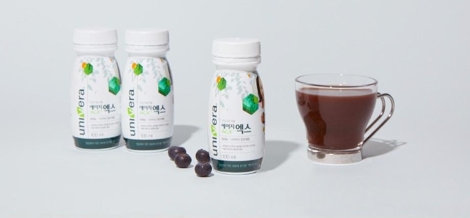 에이지엑스 100 ml 병 세 개와 짙은 자줏빛 음료를 담은 유리컵, 블랙커런트 열매가 회청색 바닥에 놓인 연출 컷