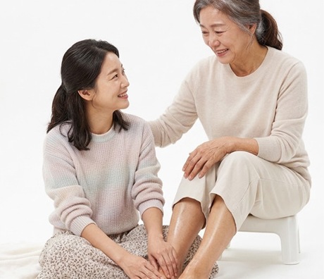흰 배경에 앉아 서로 발을 매만지며 웃는 두 여성